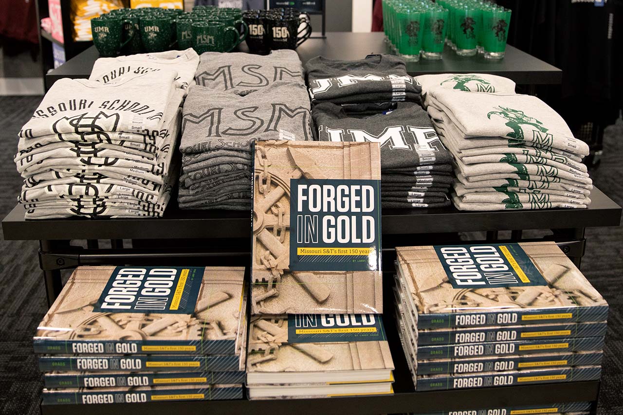 Product display table with a book, sweatshirts, tshirts, mugs, and pilsner glass.